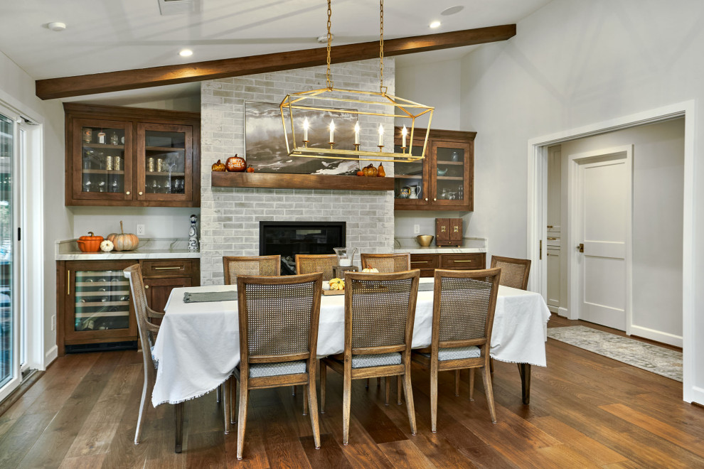 Cette image montre une grande salle à manger ouverte sur le salon traditionnelle avec un mur gris, un sol en bois brun, une cheminée standard, un manteau de cheminée en brique, un sol marron et un plafond voûté.
