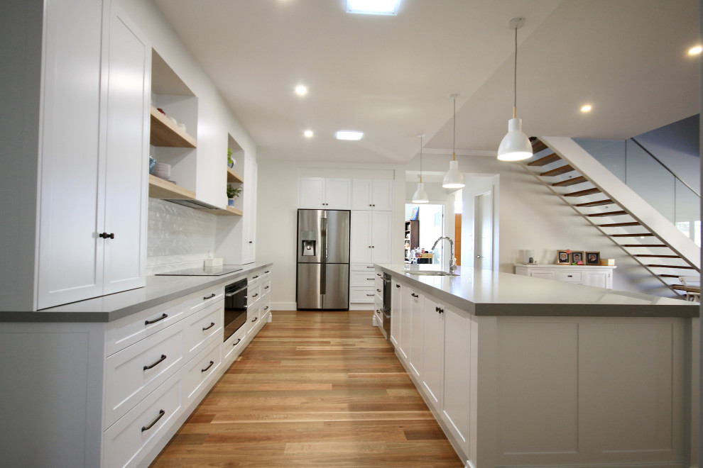 Large transitional l-shaped open plan kitchen in Sydney with an undermount sink, shaker cabinets, white cabinets, quartz benchtops, white splashback, subway tile splashback, stainless steel appliances, medium hardwood floors, with island, brown floor and grey benchtop.