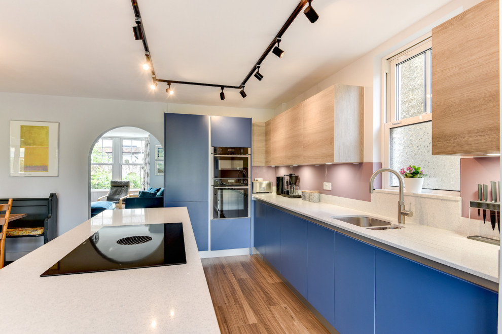 Photo of a mid-sized beach style l-shaped eat-in kitchen in Sussex with a double-bowl sink, flat-panel cabinets, blue cabinets, quartzite benchtops, multi-coloured splashback, glass sheet splashback, black appliances, dark hardwood floors, with island, brown floor and white benchtop.