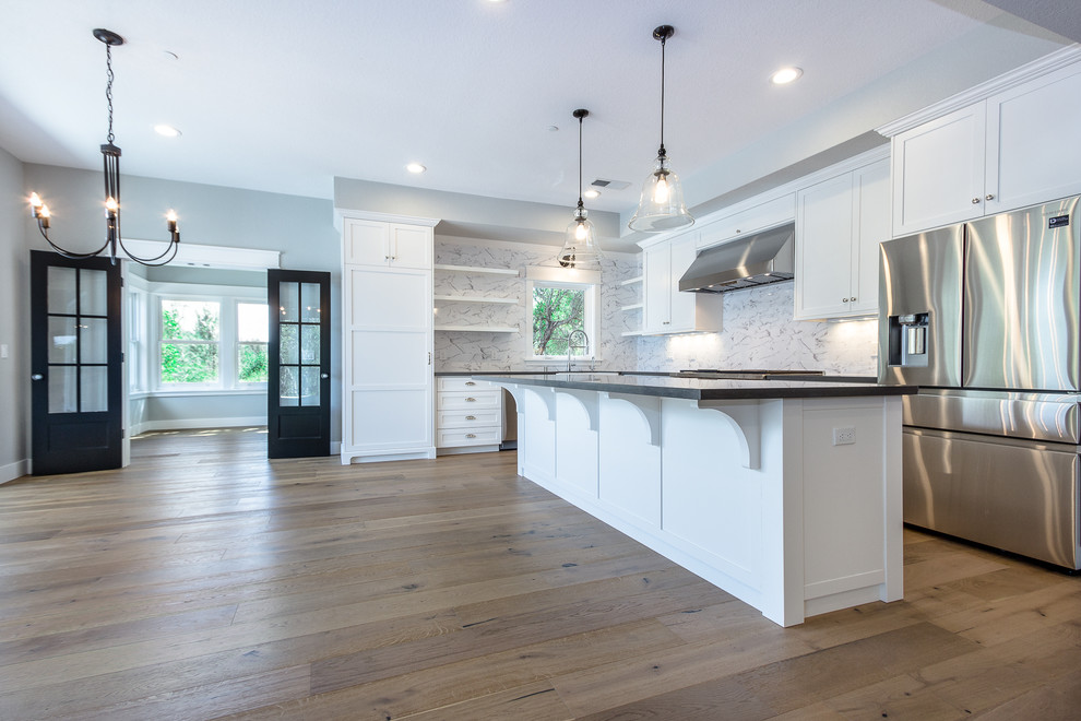 Photo of a large country l-shaped eat-in kitchen in Sacramento with a farmhouse sink, shaker cabinets, white cabinets, granite benchtops, multi-coloured splashback, stone slab splashback, stainless steel appliances, medium hardwood floors, with island, brown floor and black benchtop.