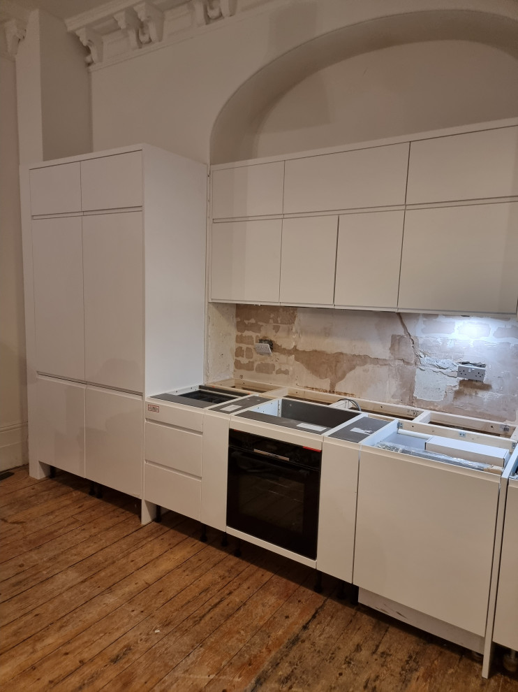 Large modern single-wall open plan kitchen in London with a drop-in sink, flat-panel cabinets, white cabinets, quartzite benchtops, white splashback, stone slab splashback, white appliances, painted wood floors and white benchtop.