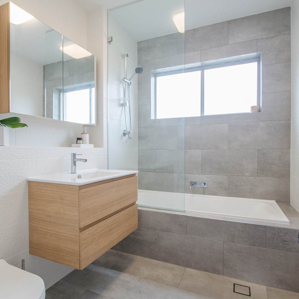 Photo of a small contemporary bathroom in Sydney with flat-panel cabinets, light wood cabinets, a drop-in tub, a shower/bathtub combo, gray tile, ceramic tile, engineered quartz benchtops, a two-piece toilet, white walls, ceramic floors and a drop-in sink.