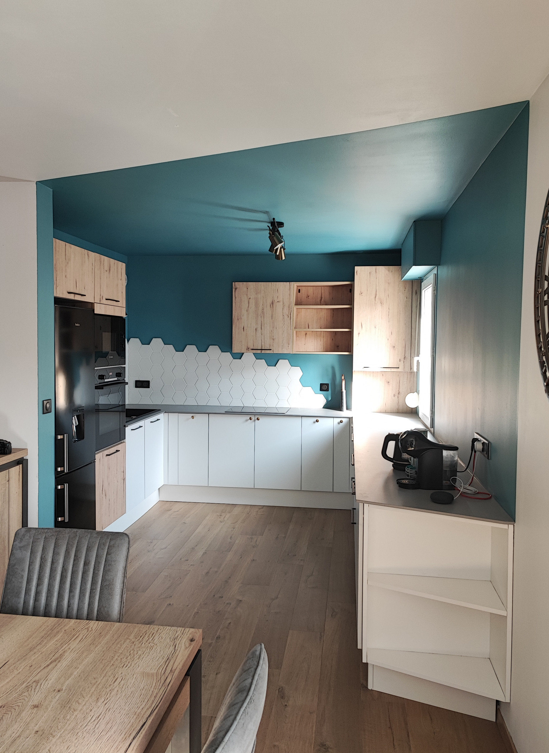 Vue d'ensemble de la cuisine en U, avec un long plan de travail à droite, pour le petit électroménager, le coin technique à gauche qui cache l'évier et la plaque en face.