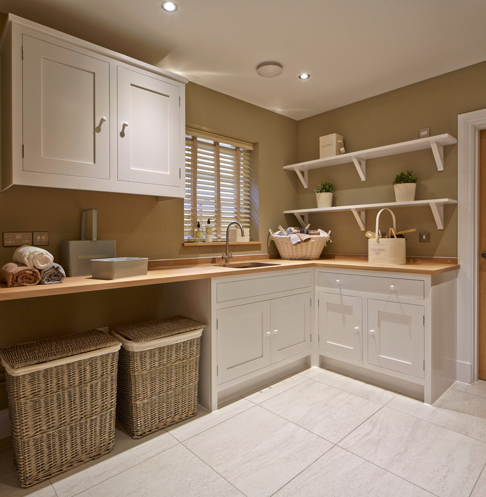 Photo of a country l-shaped utility room in Wiltshire with an undermount sink, shaker cabinets, white cabinets, wood benchtops, beige walls, grey floor and beige benchtop.