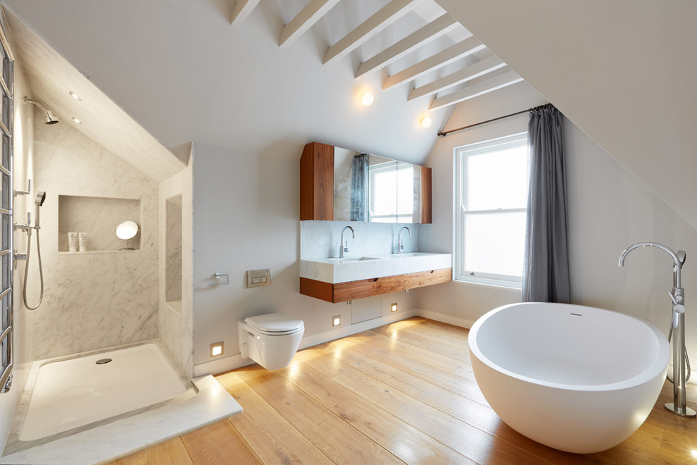 Large modern bathroom in London with flat-panel cabinets, brown cabinets, a freestanding tub, a wall-mount toilet, white walls, light hardwood floors, a wall-mount sink, marble benchtops, beige floor, an open shower and grey benchtops.