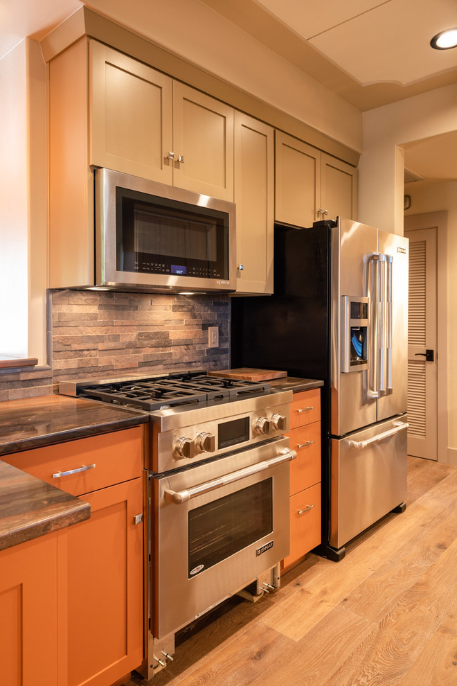 This is an example of a mid-sized arts and crafts galley eat-in kitchen in San Luis Obispo with a drop-in sink, shaker cabinets, orange cabinets, granite benchtops, multi-coloured splashback, stone tile splashback, stainless steel appliances, light hardwood floors, a peninsula, brown floor and multi-coloured benchtop.