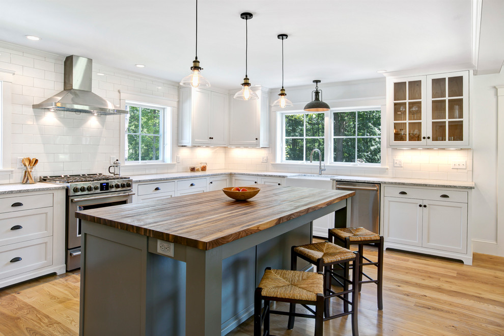 This is an example of a country l-shaped kitchen in Burlington with a farmhouse sink, glass-front cabinets, white cabinets, white splashback, subway tile splashback, stainless steel appliances, light hardwood floors, with island, beige floor, grey benchtop and granite benchtops.