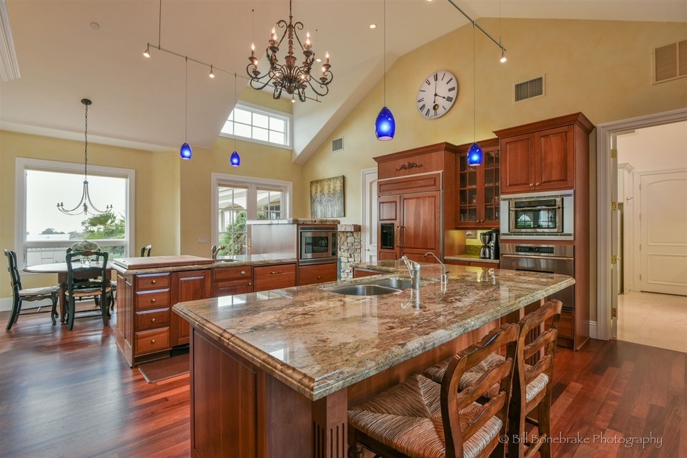 This is an example of a large traditional u-shaped eat-in kitchen in Austin with a double-bowl sink, raised-panel cabinets, medium wood cabinets, granite benchtops, beige splashback, stainless steel appliances, medium hardwood floors and multiple islands.