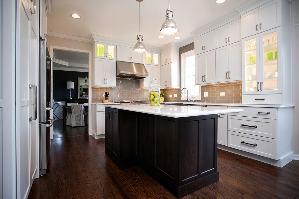 Example of a large transitional l-shaped dark wood floor enclosed kitchen design in Chicago with a farmhouse sink, recessed-panel cabinets, white cabinets, marble countertops, beige backsplash, glass tile backsplash, stainless steel appliances and an island