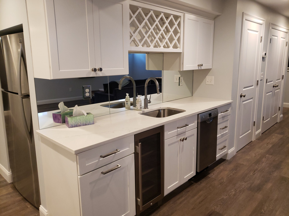 Photo of a small transitional single-wall wet bar in Boston with an undermount sink, shaker cabinets, white cabinets, quartz benchtops, mirror splashback, vinyl floors, brown floor and white benchtop.