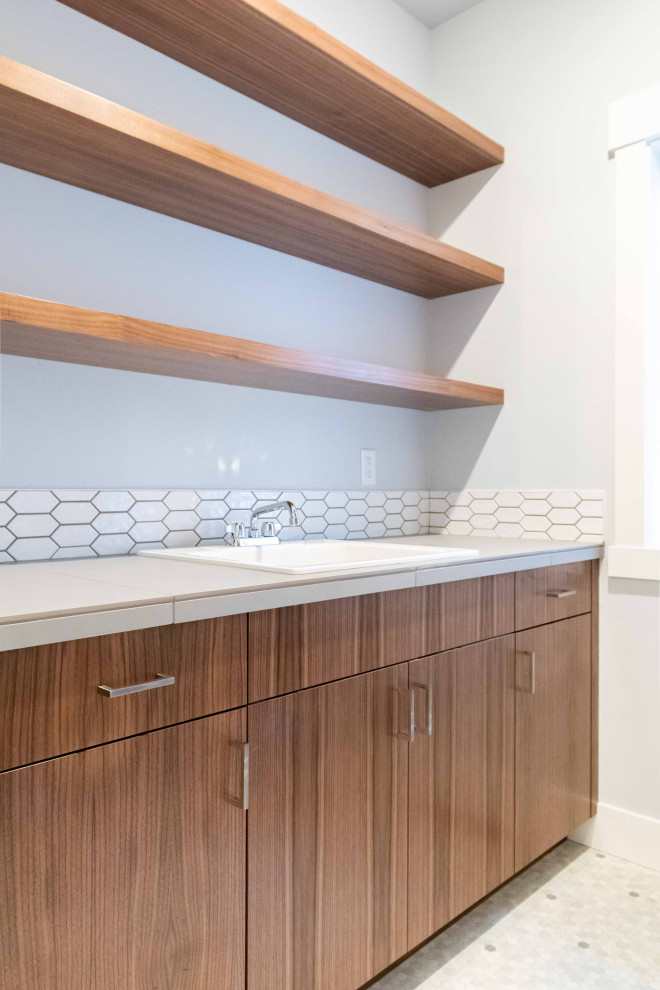 This is an example of a mid-sized modern galley dedicated laundry room in Other with a drop-in sink, flat-panel cabinets, medium wood cabinets, tile benchtops, white splashback, ceramic splashback, grey walls, vinyl floors, a side-by-side washer and dryer, grey floor and beige benchtop.