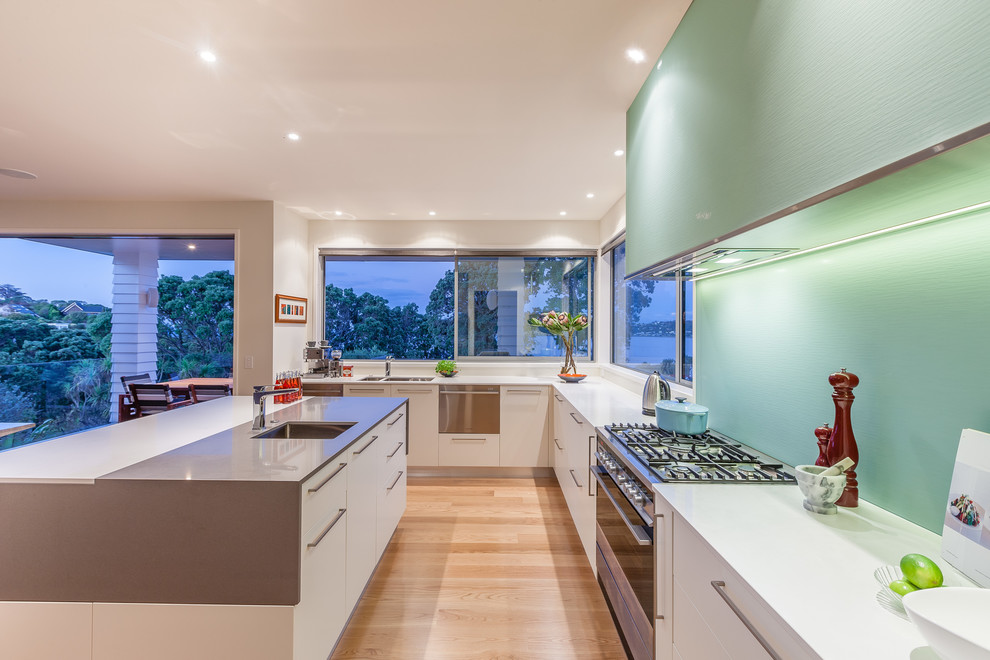 Design ideas for a mid-sized tropical l-shaped kitchen pantry in Auckland with an undermount sink, flat-panel cabinets, quartz benchtops, green splashback, glass sheet splashback, stainless steel appliances, light hardwood floors and with island.