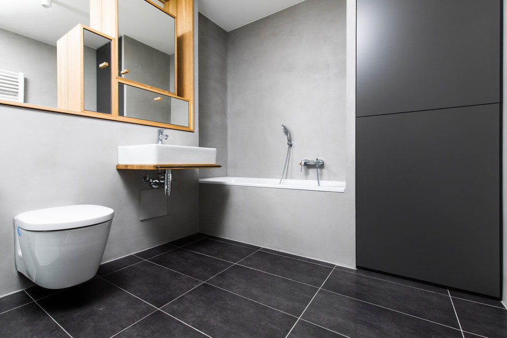 Design ideas for a mid-sized contemporary 3/4 bathroom in Munich with glass-front cabinets, a drop-in tub, a wall-mount toilet, grey walls, cement tiles, a vessel sink, wood benchtops, black floor and brown benchtops.