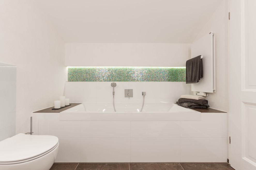Badezimmer With flächenbündigen Schrankfronten, weißen Schränken, Einbaubadewanne, Duschnische, Wandtoilette, grünen Fliesen, Glasfliesen, weißer Wandfarbe, Kalkstein, Aufsatzwaschbecken, Edelstahl-Waschbecken/Waschtisch, braunem Boden and Falttür-Duschabtrennung in Munich