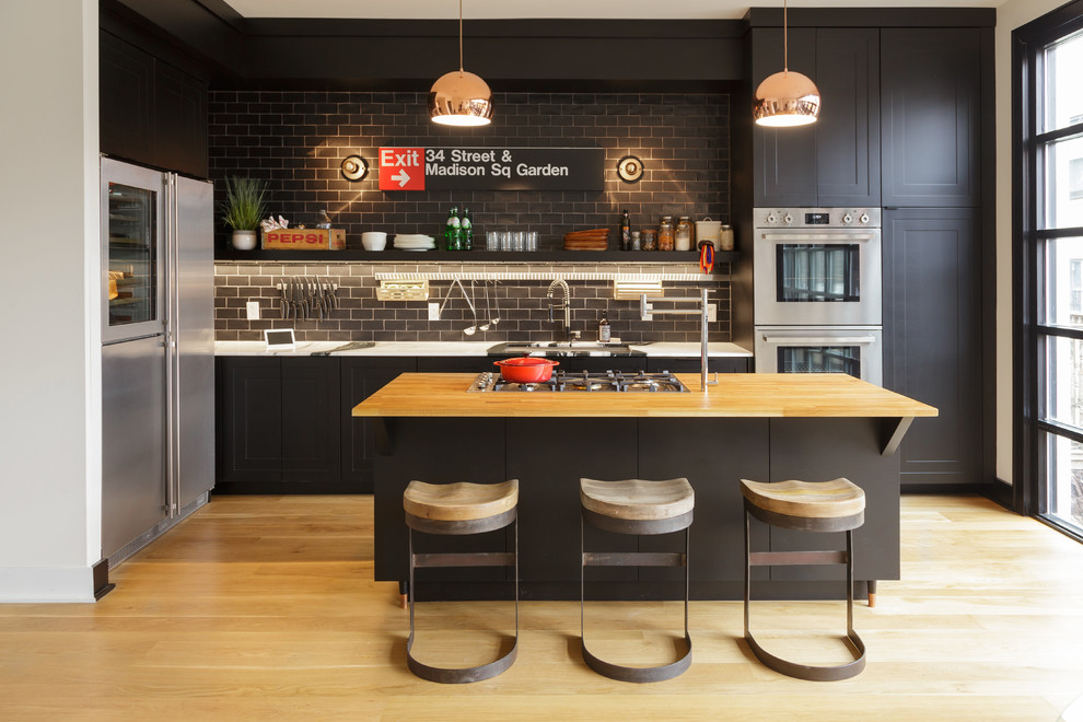 Inspiration for a mid-sized industrial galley light wood floor open concept kitchen remodel in Philadelphia with an undermount sink, shaker cabinets, black cabinets, wood countertops, gray backsplash, subway tile backsplash, stainless steel appliances and an island