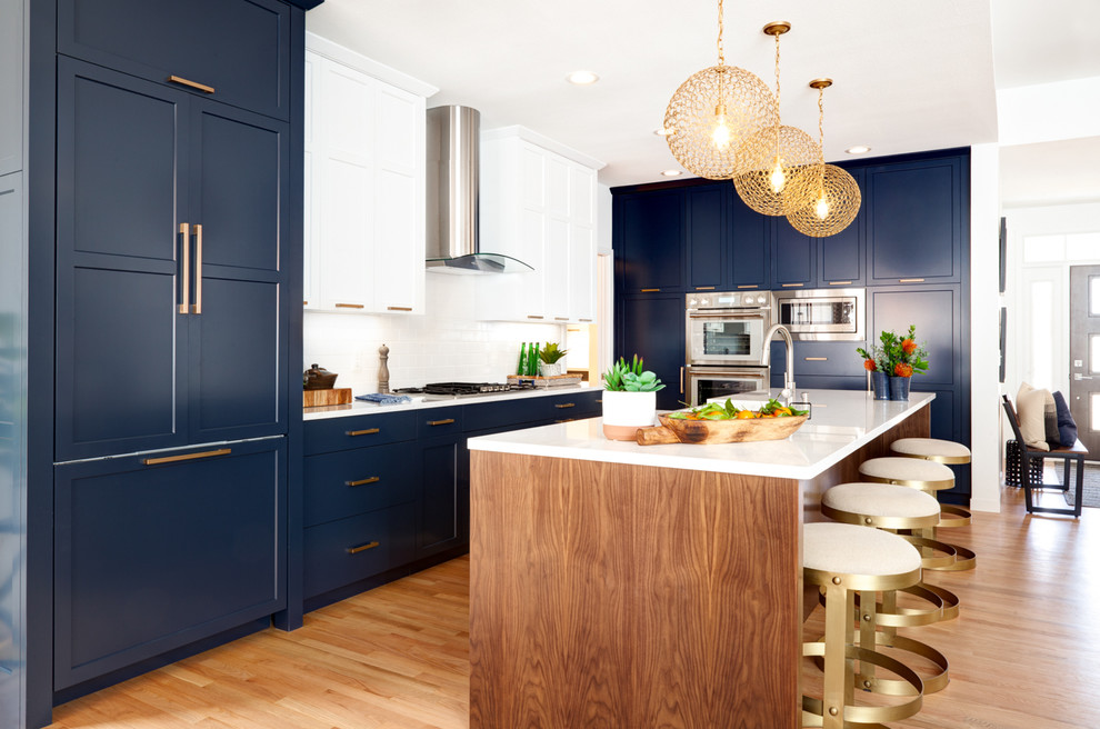 Photo of a mid-sized transitional galley kitchen in Minneapolis with blue cabinets, quartz benchtops, white splashback, porcelain splashback, stainless steel appliances, medium hardwood floors, with island, white benchtop, a farmhouse sink and recessed-panel cabinets.