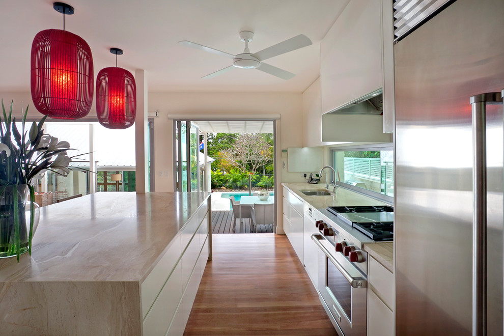 Expansive beach style galley open plan kitchen in Sunshine Coast with a double-bowl sink, flat-panel cabinets, white cabinets, marble benchtops, glass sheet splashback, stainless steel appliances, medium hardwood floors and with island.