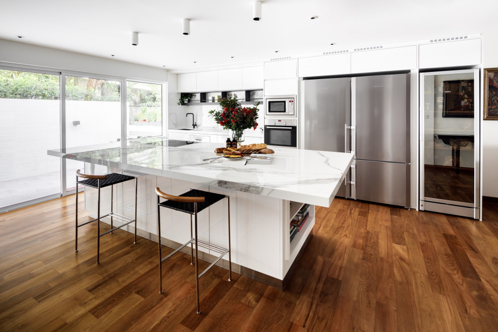 Inspiration for a large contemporary galley kitchen in Singapore with an undermount sink, flat-panel cabinets, white cabinets, white splashback, glass sheet splashback, stainless steel appliances, medium hardwood floors, with island, brown floor and white benchtop.