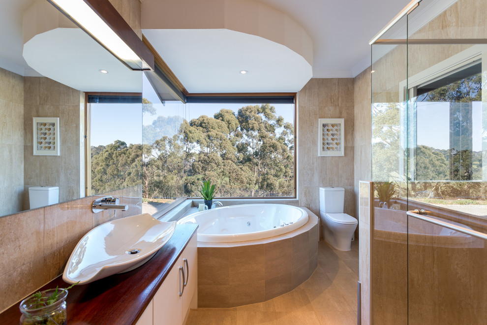 Photo of a large contemporary master bathroom in Adelaide with a vessel sink, white cabinets, beige tile, wood benchtops, a corner shower, a one-piece toilet, stone tile, a drop-in tub and flat-panel cabinets.