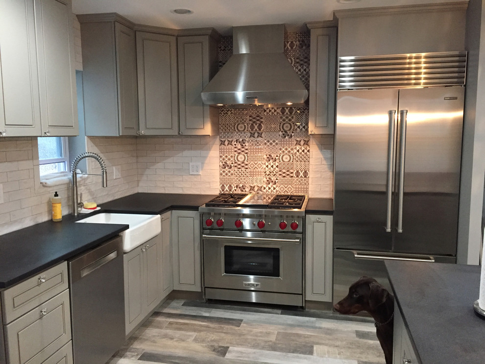 Photo of a mid-sized transitional l-shaped separate kitchen in New York with a farmhouse sink, raised-panel cabinets, grey cabinets, soapstone benchtops, grey splashback, cement tile splashback, stainless steel appliances, medium hardwood floors, with island, multi-coloured floor and black benchtop.
