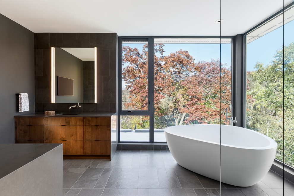 Photo of an expansive contemporary master bathroom in Minneapolis with flat-panel cabinets, brown cabinets, a freestanding tub, a curbless shower, a wall-mount toilet, black tile, porcelain tile, black walls, porcelain floors, an integrated sink, engineered quartz benchtops, grey floor, an open shower, black benchtops, an enclosed toilet, a double vanity and a built-in vanity.