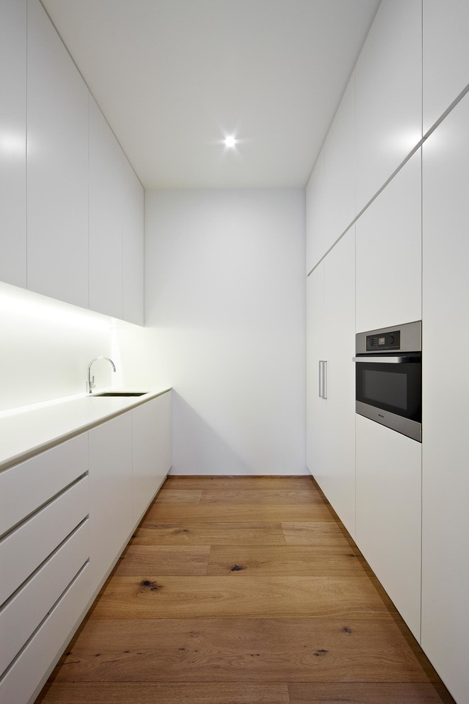 This is an example of a small scandinavian galley separate kitchen in Melbourne with flat-panel cabinets, white cabinets, panelled appliances and light hardwood floors.