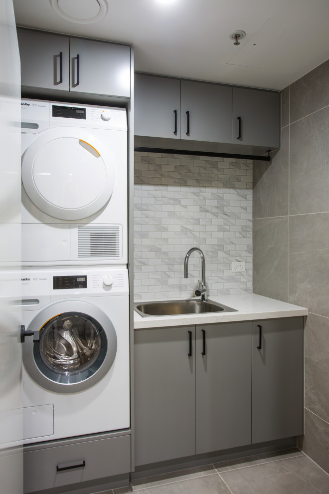 Small modern single-wall dedicated laundry room in Brisbane with a drop-in sink, flat-panel cabinets, grey cabinets, quartz benchtops, grey walls, porcelain floors, a stacked washer and dryer, grey floor and white benchtop.