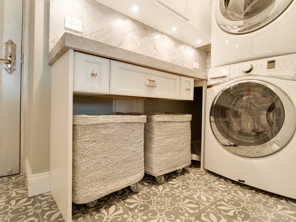 Photo of a small transitional l-shaped utility room in New York with white cabinets, marble benchtops, grey walls, concrete floors, a stacked washer and dryer, grey floor, grey benchtop and recessed-panel cabinets.