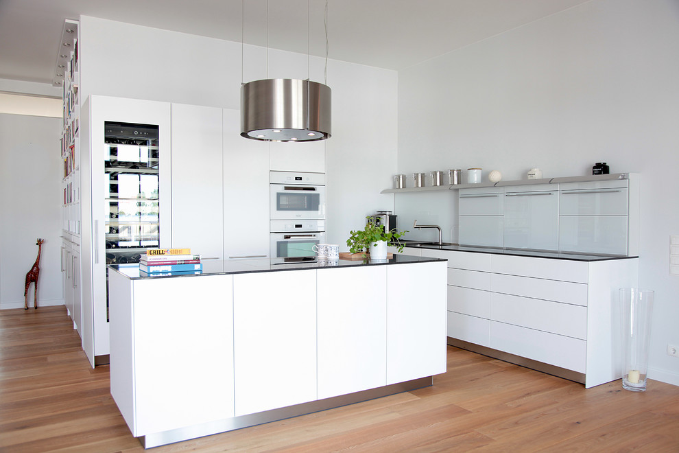 This is an example of a mid-sized contemporary l-shaped open plan kitchen in Hamburg with an integrated sink, flat-panel cabinets, white cabinets, glass benchtops, white splashback, glass sheet splashback, white appliances, medium hardwood floors, with island, brown floor and black benchtop.