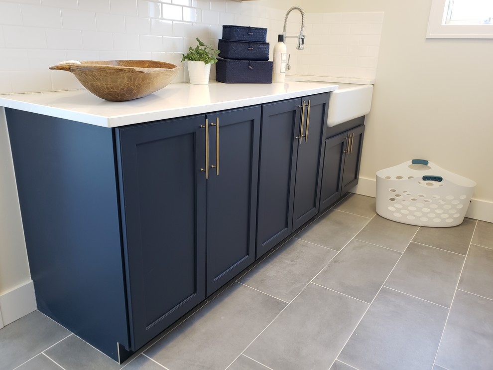 Photo of a small arts and crafts galley dedicated laundry room in Detroit with a farmhouse sink, recessed-panel cabinets, blue cabinets, quartzite benchtops, beige walls, ceramic floors, a side-by-side washer and dryer, grey floor and white benchtop.