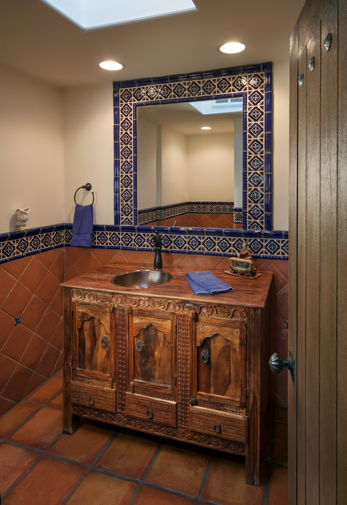 This is an example of a small mediterranean powder room in Phoenix with furniture-like cabinets, wood benchtops, blue tile, orange tile, terra-cotta floors, terra-cotta tile, beige walls, a drop-in sink, dark wood cabinets and brown benchtops.