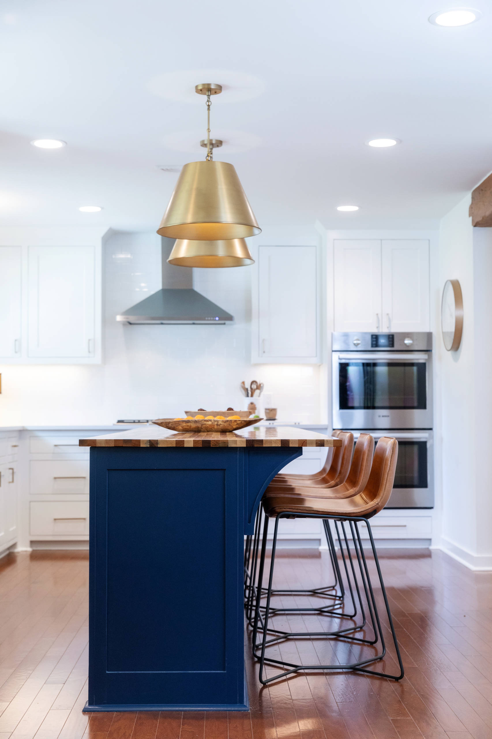 Blue kitchen island with seating. Wooden counter top. Gold pendant lights to brighten space. Gold fixtures to complement the blue paint. Stainless steel appliances. Medium hardwood flooring. White cab