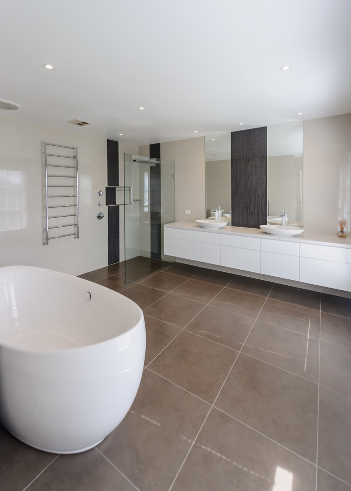 Photo of an expansive contemporary bathroom in Melbourne with a vessel sink, flat-panel cabinets, white cabinets, engineered quartz benchtops, a freestanding tub, an open shower, porcelain tile, porcelain floors, a one-piece toilet, brown tile and beige walls.