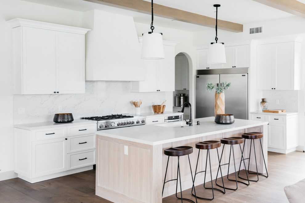 Photo of a mediterranean l-shaped kitchen in Los Angeles with an integrated sink, shaker cabinets, white cabinets, white splashback, stainless steel appliances, medium hardwood floors, with island, brown floor and white benchtop.