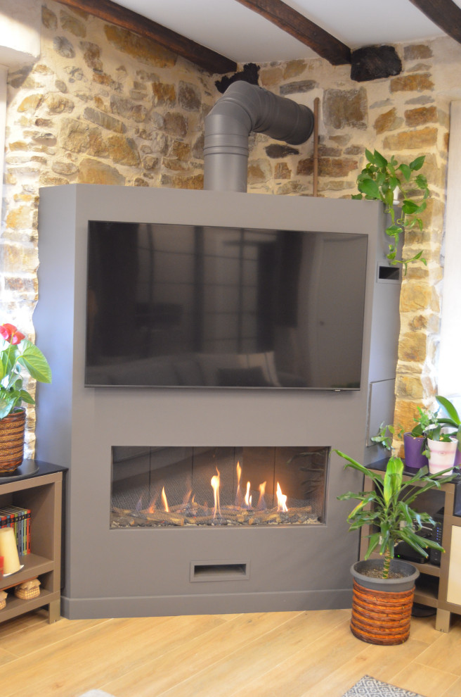 Ejemplo de salón abierto rural grande con paredes beige, suelo de madera clara, chimenea de esquina, marco de chimenea de metal, televisor en una esquina, suelo marrón, vigas vistas, piedra y alfombra