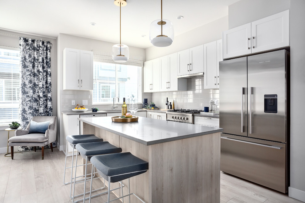 Photo of a beach style l-shaped kitchen in Vancouver with an undermount sink, shaker cabinets, white cabinets, grey splashback, stainless steel appliances, light hardwood floors, with island, grey floor and grey benchtop.