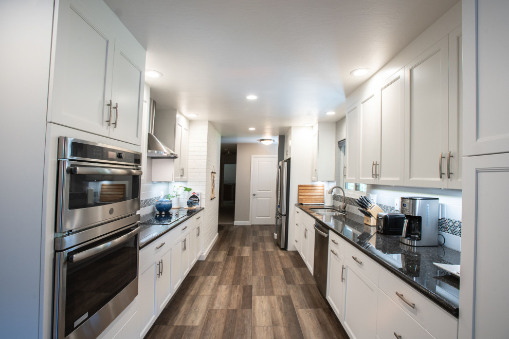 This is an example of a small contemporary galley kitchen in San Francisco with a double-bowl sink, recessed-panel cabinets, white cabinets, grey splashback, subway tile splashback, stainless steel appliances, no island, brown floor and black benchtop.