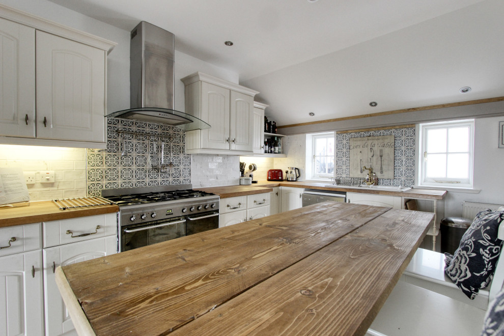 Design ideas for a mid-sized traditional galley open plan kitchen in Edinburgh with a drop-in sink, shaker cabinets, white cabinets, wood benchtops, white splashback, ceramic splashback, stainless steel appliances, porcelain floors, with island, beige floor and brown benchtop.