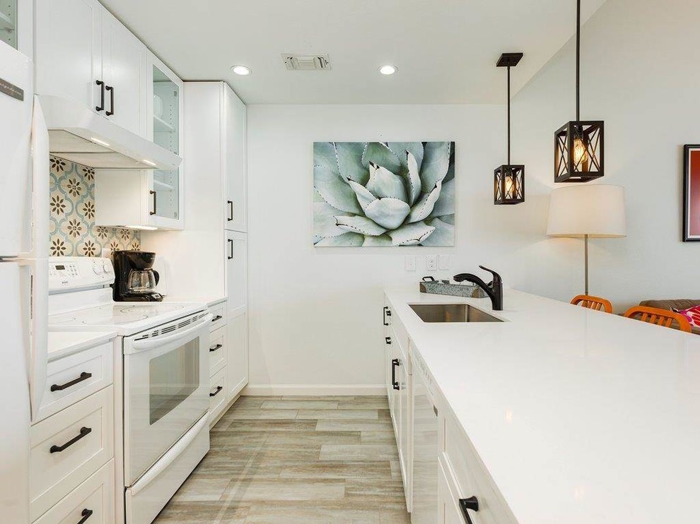 This is an example of a mid-sized beach style galley open plan kitchen in Houston with a single-bowl sink, shaker cabinets, white cabinets, quartz benchtops, multi-coloured splashback, porcelain splashback, vinyl floors, white benchtop, white appliances, no island and beige floor.