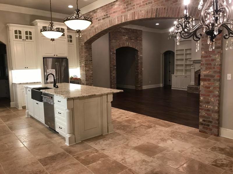 Large country single-wall open plan kitchen in New Orleans with a farmhouse sink, raised-panel cabinets, white cabinets, granite benchtops, stainless steel appliances, travertine floors, with island and brown floor.