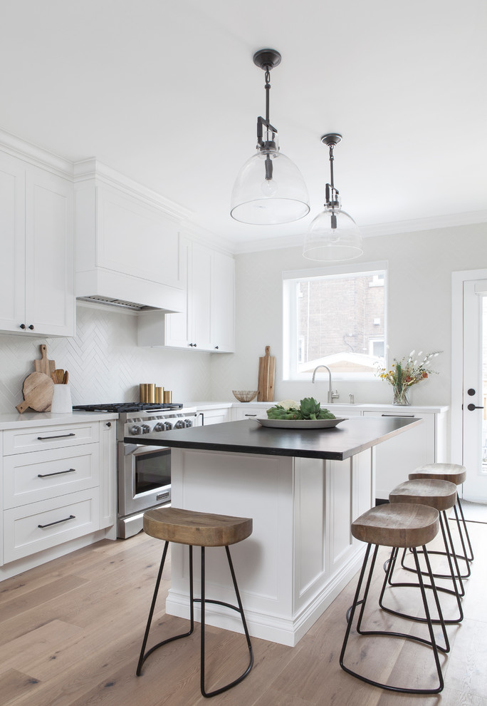Example of a small transitional l-shaped light wood floor and brown floor kitchen pantry design in Toronto with a double-bowl sink, recessed-panel cabinets, white cabinets, soapstone countertops, white backsplash, porcelain backsplash, stainless steel appliances, an island and black countertops