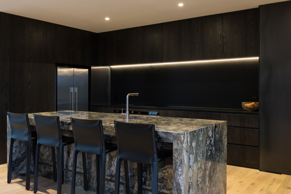 Inspiration for a mid-sized industrial galley eat-in kitchen in Auckland with a single-bowl sink, flat-panel cabinets, dark wood cabinets, marble benchtops, black splashback, slate splashback, stainless steel appliances, light hardwood floors, with island, brown floor and black benchtop.