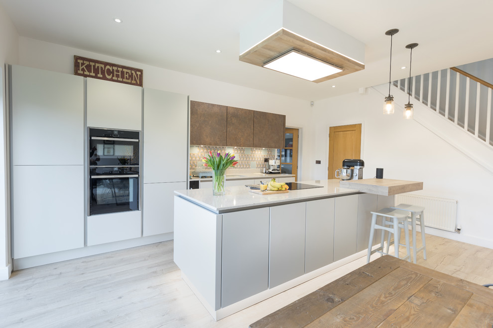 This is an example of a mid-sized modern single-wall open plan kitchen in Hampshire with an undermount sink, flat-panel cabinets, white cabinets, quartzite benchtops, metallic splashback, panelled appliances, light hardwood floors and with island.
