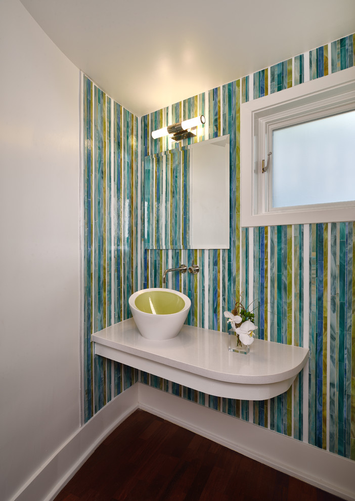 Photo of a small contemporary powder room in San Diego with a one-piece toilet, multi-coloured tile, glass tile, white walls, medium hardwood floors, a vessel sink, solid surface benchtops, brown floor, white cabinets, white benchtops and a floating vanity.