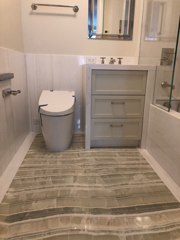 Photo of a mid-sized transitional master bathroom in New York with recessed-panel cabinets, beige cabinets, an undermount tub, a shower/bathtub combo, a one-piece toilet, white tile, stone slab, white walls, marble floors, an undermount sink, onyx benchtops, beige floor, a hinged shower door, white benchtops, a niche, a single vanity and a built-in vanity.