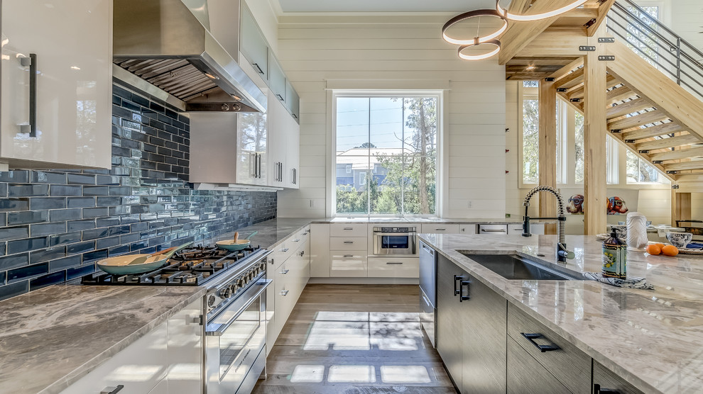 Expansive beach style u-shaped open plan kitchen in Miami with an undermount sink, flat-panel cabinets, white cabinets, subway tile splashback, stainless steel appliances, medium hardwood floors, with island, brown floor, marble benchtops, blue splashback and multi-coloured benchtop.