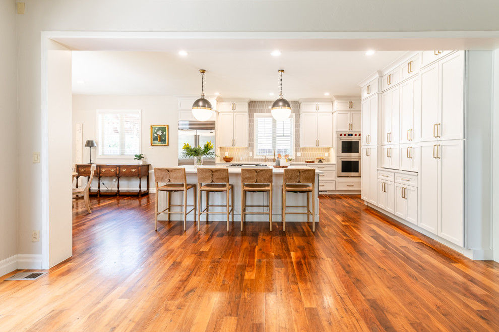 Example of a large transitional l-shaped dark wood floor and brown floor open concept kitchen design in Denver with a farmhouse sink, shaker cabinets, beige cabinets, quartz countertops, beige backsplash, ceramic backsplash, stainless steel appliances, an island and white countertops