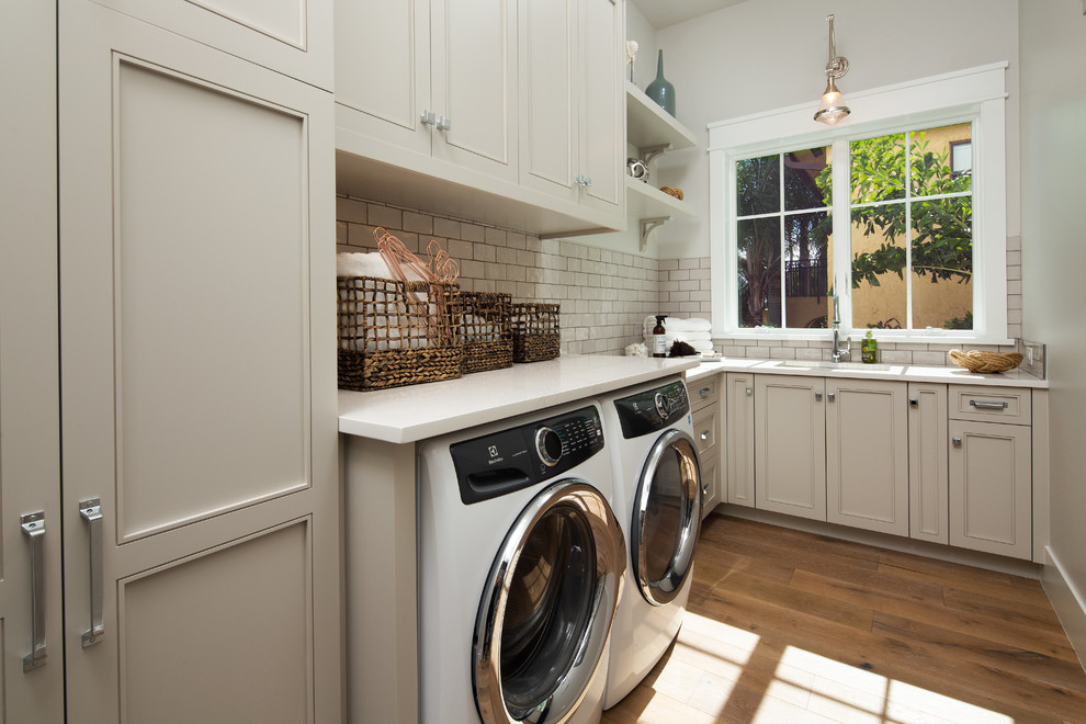 Inspiration for a beach style l-shaped dedicated laundry room in Miami with an undermount sink, recessed-panel cabinets, beige cabinets, grey walls, light hardwood floors, a side-by-side washer and dryer and white benchtop.
