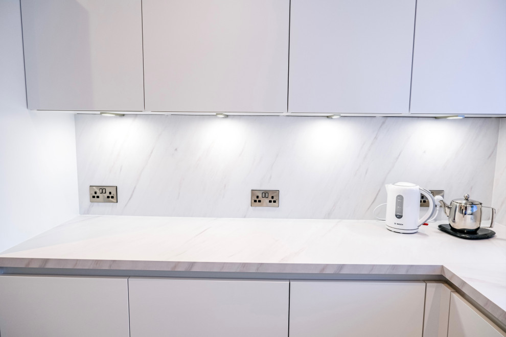 Cette image montre une cuisine américaine parallèle minimaliste de taille moyenne avec des portes de placard blanches, une crédence blanche, une crédence en feuille de verre, un électroménager noir, aucun îlot et un plan de travail blanc.