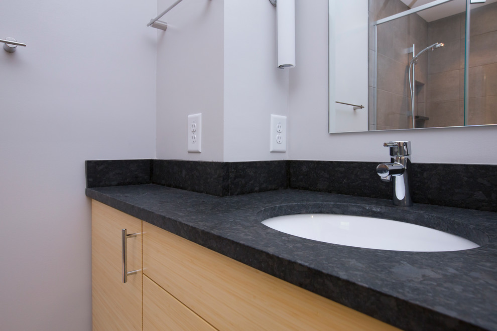 This is an example of a mid-sized contemporary master bathroom in Raleigh with flat-panel cabinets, light wood cabinets, a corner shower, a one-piece toilet, gray tile, porcelain tile, grey walls, porcelain floors, an undermount sink, granite benchtops, grey floor, a hinged shower door and black benchtops.